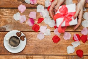 50635298 - valentines day gift in white box and female hands and petals on wooden background with cup of coffee